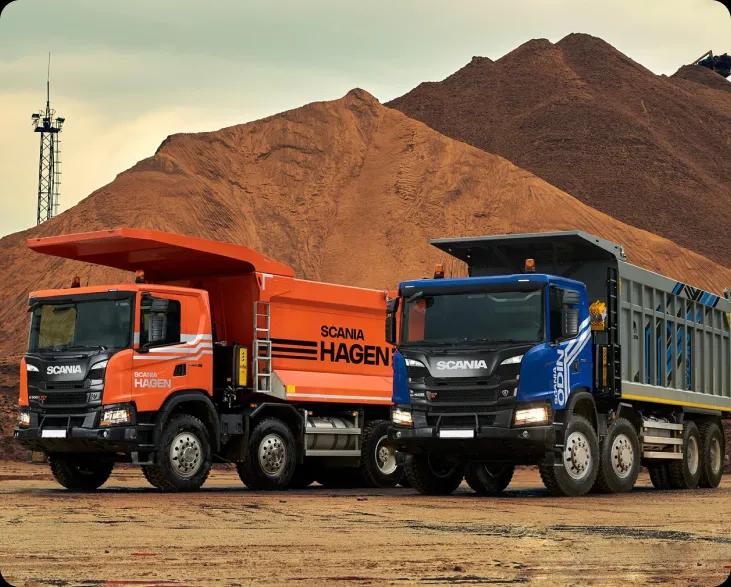 Два самосвала Scania, один оранжевый, а другой синий, стоят перед кучей земли
