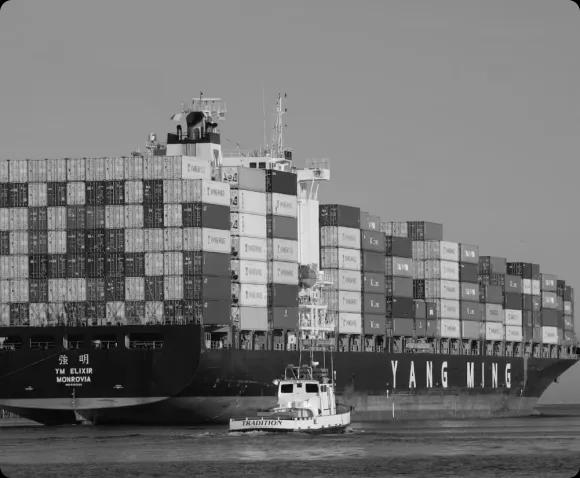 Черно-белое фото большого контейнеровоза, полного грузовых контейнеров, и малого судна на переднем плане, демонстрирующее масштабы морской логистики.