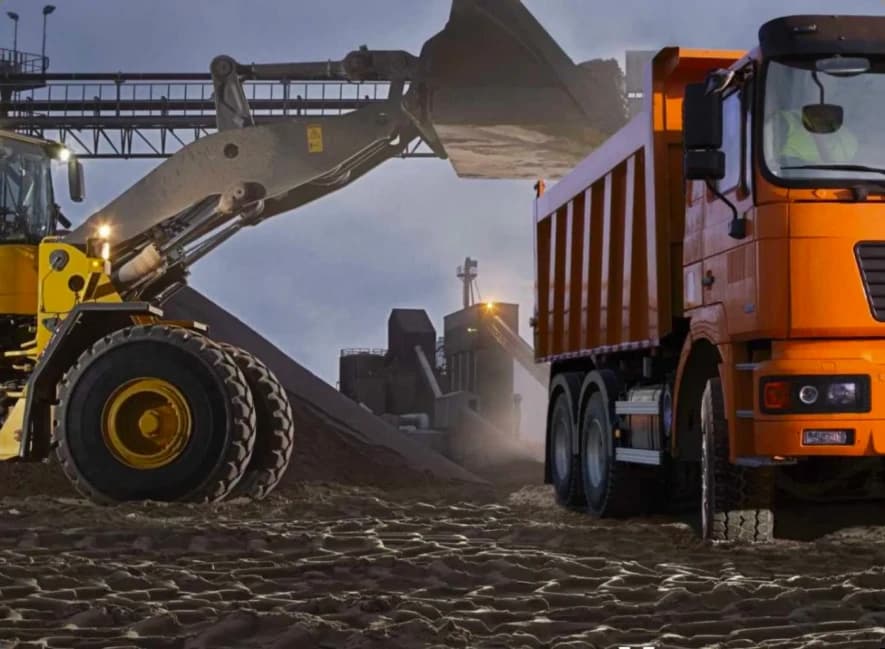 Колёсный погрузчик выгружает песок в кузов оранжевого грузовика на строительной площадке.