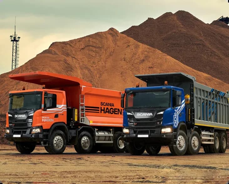 Два самосвала Scania, один оранжевый, а другой синий, стоят перед кучей земли.